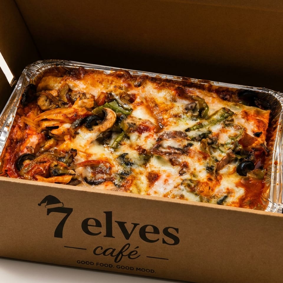 Baked vegetable lasagna topped with melted cheese in a foil tray inside a 7elves Café takeout box.
